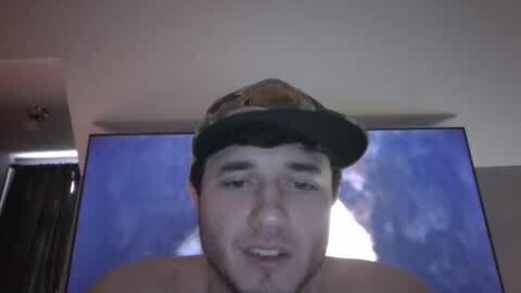 Media: Video of a shirtless young man with a baseball cap, watching a TV screen showing a football game. The room is dimly lit, with a ceiling fan visible.