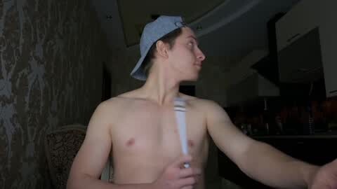 Media: Video of a shirtless young man with a slim build, wearing a backward blue cap, standing indoors, with a white strap across his chest. Background features a patterned wall and dimly lit kitchen.