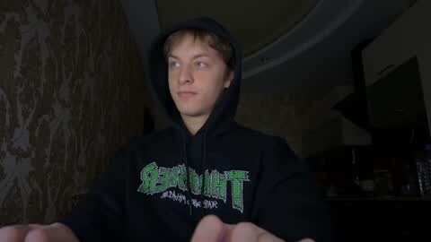 Media: Video of a young, fair-skinned, light-haired man with a serious expression, wearing a black hoodie with green text. He stands in a dimly lit room with peeling wallpaper and a ceiling fan.