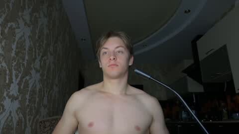Media: Video of a shirtless, light-skinned, young man with short blond hair, standing indoors with a dimly lit background featuring a patterned wall and modern lighting fixtures.