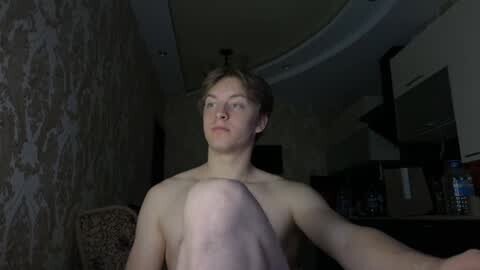 Media: Video of a shirtless, slender, fair-skinned, young man with short, light brown hair sitting on a couch in a dimly lit room with patterned wallpaper, a TV, and a dark staircase in the background.