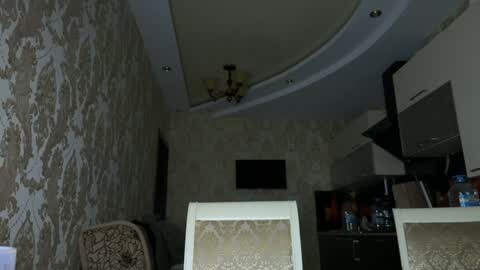 Media: A video of a dimly lit, vintage-style room with beige floral wallpaper, a curved white ceiling, and a flat-screen TV mounted above a dark wood cabinet. The room features a patterned chair and a table with water bottles.