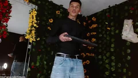 Media: Video of a muscular man with short dark hair in a black long-sleeve shirt and light blue jeans, holding a black object, standing in a room with vibrant floral wallpaper and a white statue.