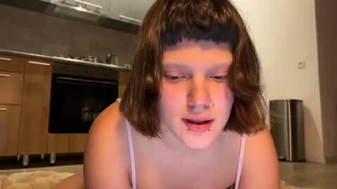 Video of a young woman with shoulder-length brown hair, wearing a white spaghetti strap top, sitting in a modern kitchen with light wooden cabinets and stainless steel appliances.