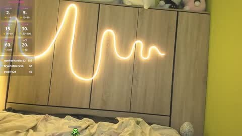 Media: Video of a bedroom with a wooden headboard displaying a glowing, animated \"U\" graphic. A white bed with crumpled beige sheets is in the foreground. The room has a yellow wall and a digital clock displaying 2:05 AM.