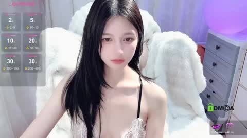 Media: Video of a slim Asian woman with long black hair, wearing a lacy white bra, posing on a white fur blanket in a bedroom with purple lighting.