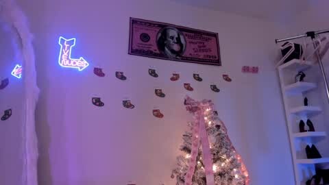 Media: A video of a cozy, festive room with a lit-up Christmas tree adorned with snowflake ornaments, a neon \"Merry\" sign, and a Benjamin Franklin dollar bill on the white wall.