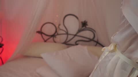 Media: Video of a woman in a sheer, white nightgown with a black, ornate metal frame draped over her bed. Soft, warm lighting creates a romantic, intimate atmosphere.
