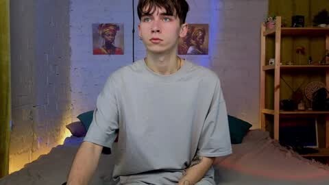 Media: Video of a young, slim, fair-skinned male with short dark hair, wearing a loose gray t-shirt, sitting on a bed in a dimly lit room with wooden shelves and colorful wall art.