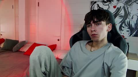 Media: Video of a young, slim, light-skinned man with dark hair, wearing a grey shirt and sweatpants, seated on a gaming chair in a modern bedroom with red lighting, white walls, and minimalist decor.