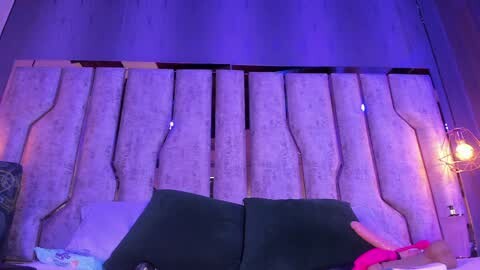 Media: Video of a dimly lit bedroom with a modern, textured, beige headboard. A dark green sofa with white pillows is in the foreground. A pink blanket and a lamp with a clear glass shade are visible on the right.