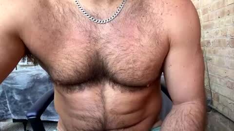 Media: Video of a muscular, shirtless man with a full chest of dark hair, wearing a silver chain necklace, indoors near a brick wall.