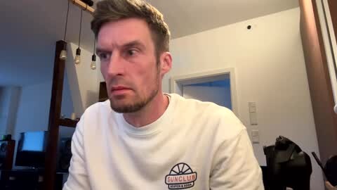 Media: Video of a bearded, light-skinned, middle-aged man with short brown hair, wearing a white \"Gong Show\" t-shirt, sitting in a dimly-lit, cluttered room with exposed bulbs and wooden furniture.