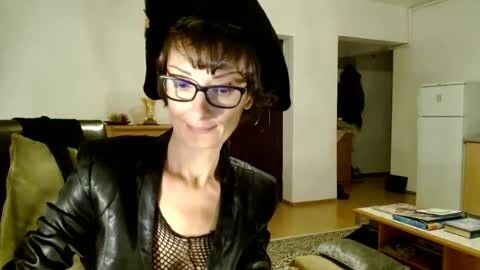 Media: Video of a woman with short dark hair, glasses, and black leather jacket, sitting on a couch in a simple living room with a wooden floor.