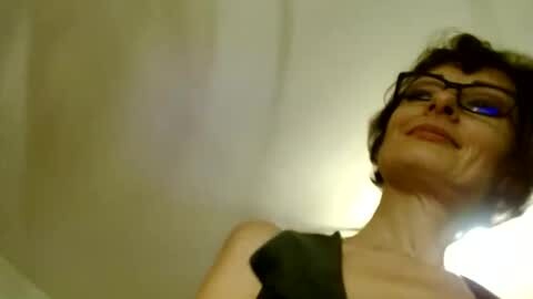 Media: A video of a woman with short, dark hair, wearing black-rimmed glasses, and a black top, smiling slightly, against a blurred background.
