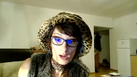 Media: Video of a white woman with short brown hair, wearing glasses, a leopard print hat, and a fur vest, speaking in a dimly-lit, sparsely furnished room.