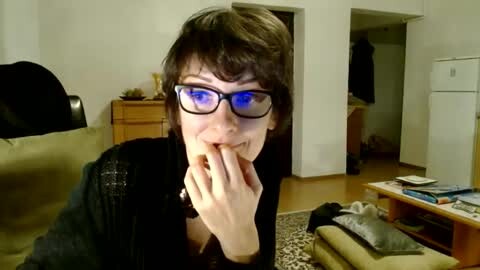 Media: A video of a person with short brown hair, wearing glasses, a black jacket, and a black shirt, sitting on a beige couch, looking at a camera, in a cluttered living room.