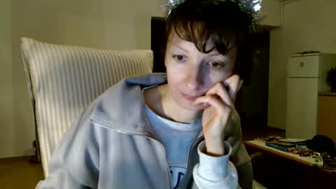 Media: A video of a young person with short brown hair, wearing a light gray hoodie, sitting in a chair with a white cushion, indoors with a white fridge and a cluttered table in the background.