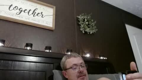 Media: Video of a man with glasses, short hair, and a beard, lying on a dark wooden couch, with a framed quote \"together all the way\" and a decorative vine on the wall.