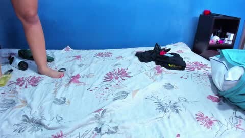 Media: Video of a messy bedroom with a blue wall, a person's bare legs stepping onto a floral bedspread, scattered clothes, and a black bag.