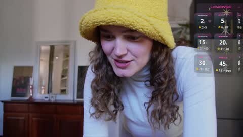 Media: Video of a curly-haired woman in a yellow beanie, white top, smiling in a cozy kitchen with a digital weather overlay.
