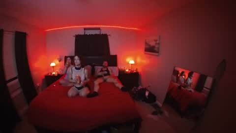 Media: A dimly-lit bedroom with red lighting, featuring a couple on a bed with a red comforter, a framed photo, and a mirror reflecting them.