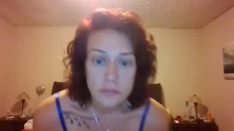 Media: Video of a young woman with curly red hair, fair skin, and blue eyes, wearing a blue tank top, sitting on a bed with beige walls, a framed picture, and a lamp on a nightstand.