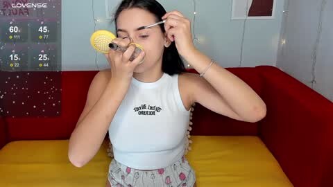 Media: Video of a young woman with long black hair, wearing a white tank top with \"poodle\" print, playing with a yellow microphone, seated on a yellow couch in a room with red and white walls.