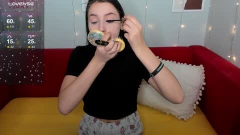 Media: Video of a young woman with fair skin, long dark hair, wearing a black t-shirt and patterned pajama pants, eating a donut and applying mascara in a cozy, red couch-filled room with star-themed wall decor.