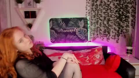Media: A video shows a red-haired woman in a black top, lounging on a red couch with a fern-patterned pillow. A black wall-mounted screen displaying \"Jungle\" and green lighting creates a modern, cozy atmosphere.