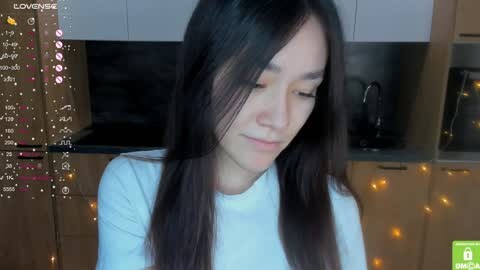 Media: A video of a young Asian woman with long, straight black hair and fair skin, wearing a white top, sitting in a modern kitchen with wooden cabinets and string lights.