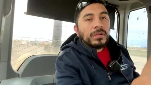 Media: Video of a bearded man with short dark hair, wearing a navy-blue uniform jacket over a red shirt, sitting in a bus. He holds a microphone, looking out the window at a cloudy sky and distant buildings.