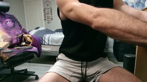 Media: Video of a muscular, shirtless man in beige shorts sitting on a chair in a cluttered bedroom. A purple blanket with a large eye design is draped over a desk, and a dreamcatcher hangs on the wall.