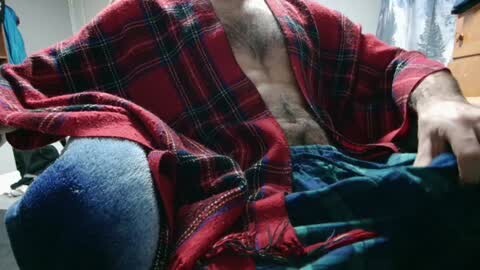 Media: Video of a shirtless man with a hairy chest, wearing a red plaid blanket and blue jeans, sitting on a bed in a dimly lit room.