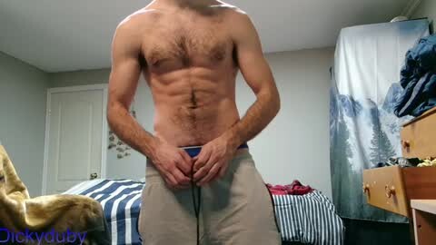 Media: Video of a shirtless, muscular man with a light tan and hairy chest, adjusting his beige shorts in a bedroom with a blue-striped bed, wooden dresser, and a forest mural on the wall.