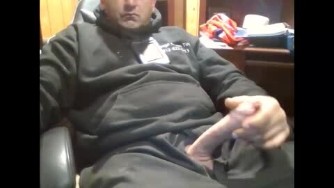 Media: A video of a middle-aged man with light skin, wearing a black hoodie with \"Pittsburgh Steelers\" printed on it, sitting in a chair, holding a large, erect penis, indoors with wooden furniture and a cluttered background.