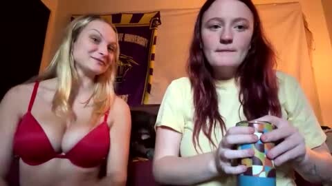 Media: Video of two women with long hair; one blonde in a red bra, the other brunette in a yellow shirt, holding a colorful mug, indoors with a purple and yellow banner in the background.