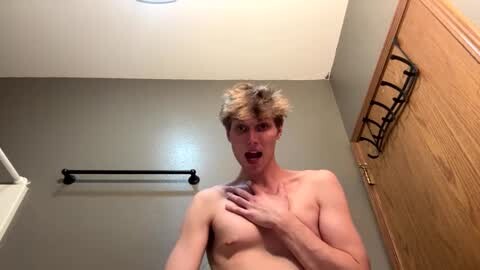 Media: Video of a shirtless, fit, fair-skinned young man with short, light brown hair, mouth open, hands on his chest, standing in a bathroom with beige walls, wooden door, and towel rack.