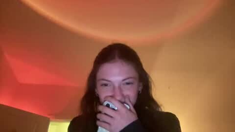 Media: Video of a young woman with long brown hair, wearing a black jacket, holding a cigarette, smiling. Background features a dim, warm-toned room with a circular red light above.
