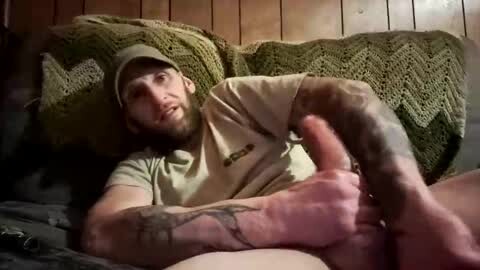 Media: Video of a bearded man with tattoos, wearing a cap and green shirt, lying on a green knitted blanket, giving a thumbs-up gesture, wooden-paneled background.