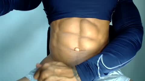 Media: Video of a muscular, dark-skinned man's toned abdomen, wearing a navy blue long-sleeve shirt, with a light-skinned hand touching his abs. Background is a blurred, neutral-toned wall.