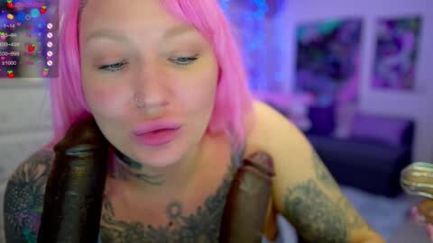 Media: Video of a tattooed, pale-skinned woman with pink hair, wearing a harness, in a dimly-lit room with purple lighting, blurred background.