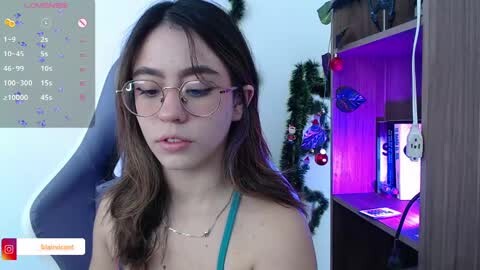 Media: Video of a young woman with long brown hair, wearing glasses and a turquoise top, sitting in a chair. Background includes a calendar, a decorated tree, and a computer monitor.
