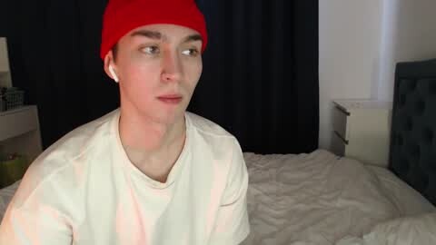 Media: Video of a young man with fair skin, wearing a red beanie, white T-shirt, and white earbuds, sitting on a bed with white sheets, against a dark curtain background.