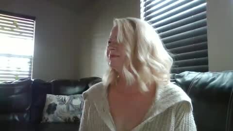 Media: Video of a blonde woman with shoulder-length hair, wearing a white robe, standing in a dimly lit living room with black leather couch and closed blinds.