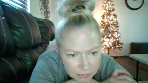 A video of a blonde woman with a high bun, wearing a grey shirt, lying face-down on a dark leather couch. A lit Christmas tree with colorful ornaments is in the background.