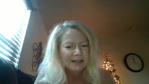 Media: A video of a middle-aged blonde woman with fair skin, medium build, and medium breasts, smiling while adjusting her hair. She has a black tattoo on her upper arm. The room features a clock and a chandelier.
