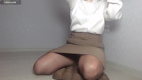Media: Video of a woman kneeling, wearing a white blouse and a tan skirt, with her legs slightly spread, in a plain, white-walled room.
