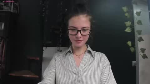 Media: Video of a young woman with glasses, light skin, and brown hair in a messy bun, wearing a striped shirt, sitting at a desk in a dimly lit room with dark walls and green ivy wallpaper.