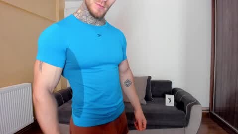 Media: Video of a muscular, bearded man in a tight blue T-shirt and brown pants, with a tattoo on his left arm, standing in a modern living room with beige walls, a grey couch, and a radiator.
